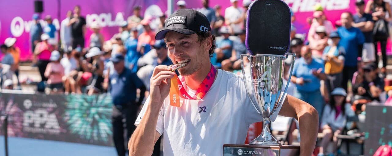 Connor Garnett biting a gold medal and holding the men's singles trophy.